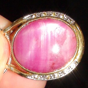 May include: An oval-shaped cabochon pink gemstone set in a gold-toned metal pendant frame. The setting features a border of small, clear, sparkling stones surrounding the central pink stone, which displays natural vertical striations and inclusions.