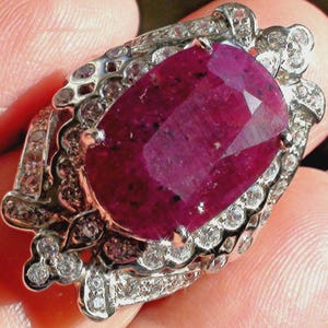 May include: A silver ring with a large, oval-cut ruby gemstone. The ruby is a deep red colour and is surrounded by smaller, clear gemstones. The ring has an ornate, detailed design.