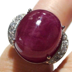 May include: A large, oval-shaped ruby ring. The ruby is a deep, rich red color and is the focal point of the ring. The ring's band is silver-colored and features a row of small, clear gemstones on each side of the ruby. The ring is shown on a finger.