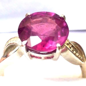 Peut inclure: Gros plan sur une bague ornée d'une grosse pierre précieuse rose foncé de forme ovale. La pierre est sertie dans un anneau argenté avec des détails décoratifs. La bague est présentée sur fond blanc, mettant en valeur la couleur vive de la pierre.