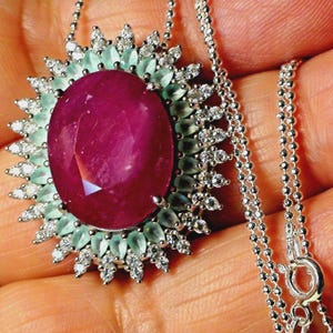 Può includere: Una collana d'argento con un grande rubino ovale come elemento centrale. Il rubino è circondato da un alone di pietre preziose verde chiaro più piccole e diamanti bianchi scintillanti. La catena è in stile delicato e perlato.