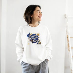 Può includere: Felpa bianca con la scritta "Dedication, Confidence, leave no space for Failure" in blu e oro. La grafica include montagne, un sole e un design a bussola. Stile casual.