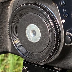 May include: A black Nikon D5100 digital SLR camera with a lens and strap, mounted on a tripod. The camera has a red accent on the top and the Nikon logo is visible. The background is blurred green grass.