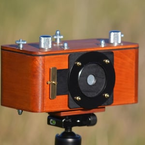May include: A rectangular wooden camera body with a polished finish, featuring a black circular lens mount, brass hardware, and silver-toned metal knobs on the top panel. The device is mounted on a black tripod head, showcasing a functional design.