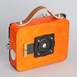 Peut inclure: Un appareil photo en bois avec une poignée en cuir et un objectif noir. L'appareil photo est orange et a un objectif noir et argent.