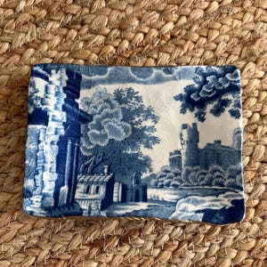 May include: Rectangular blue and white ceramic dish with a landscape scene. The design features buildings, trees, and a castle. The dish is placed on a woven, textured surface.