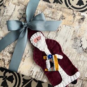 May include: A Christmas-themed gift wrapped in birch-patterned paper and tied with a light blue ribbon. A felt Santa Claus ornament in burgundy, white, and yellow is attached to the gift, adding a festive touch. The background features a patterned surface.