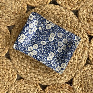 May include: A rectangular ceramic dish featuring a deep blue background with a repeating white floral pattern of small blossoms and thin stems. The dish has slightly raised edges and a smooth, glossy finish, showcasing a classic botanical design.