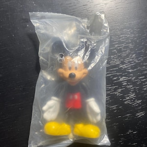 May include: A Mickey Mouse figurine in a clear plastic bag. The figure has a black body, red shorts, yellow shoes, white gloves, and a tan face. The figurine is a collectible item.