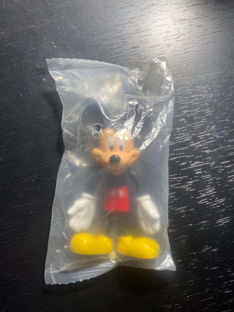 May include: A Mickey Mouse figurine in a clear plastic bag. The figure has a black body, red shorts, yellow shoes, white gloves, and a tan face. The figurine is a collectible item.