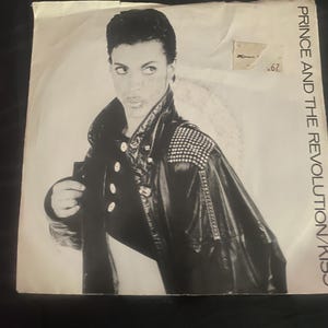 May include: Black and white album cover featuring Prince in a leather jacket with studded shoulders. The album title "Prince and The Revolution/Kiss" is printed vertically on the right side. A price tag is visible in the upper right corner.