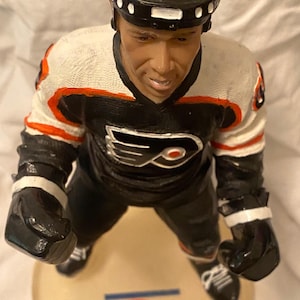 May include: A figurine of a hockey player in a black and white uniform with orange accents. The player wears a black helmet and gloves. The base of the figurine has the text "browndaub4less.com" and a logo.