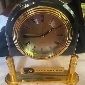 May include: A gold-toned quartz clock with a black arched frame. The clock face has Roman numerals and the words "Quartz". The base features a plaque with the Philadelphia Flyers logo and team name.