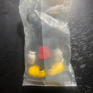 May include: A black Mickey Mouse figurine in a clear plastic bag. The figurine has yellow shoes, white gloves, and red shorts. The figure is on a dark surface.