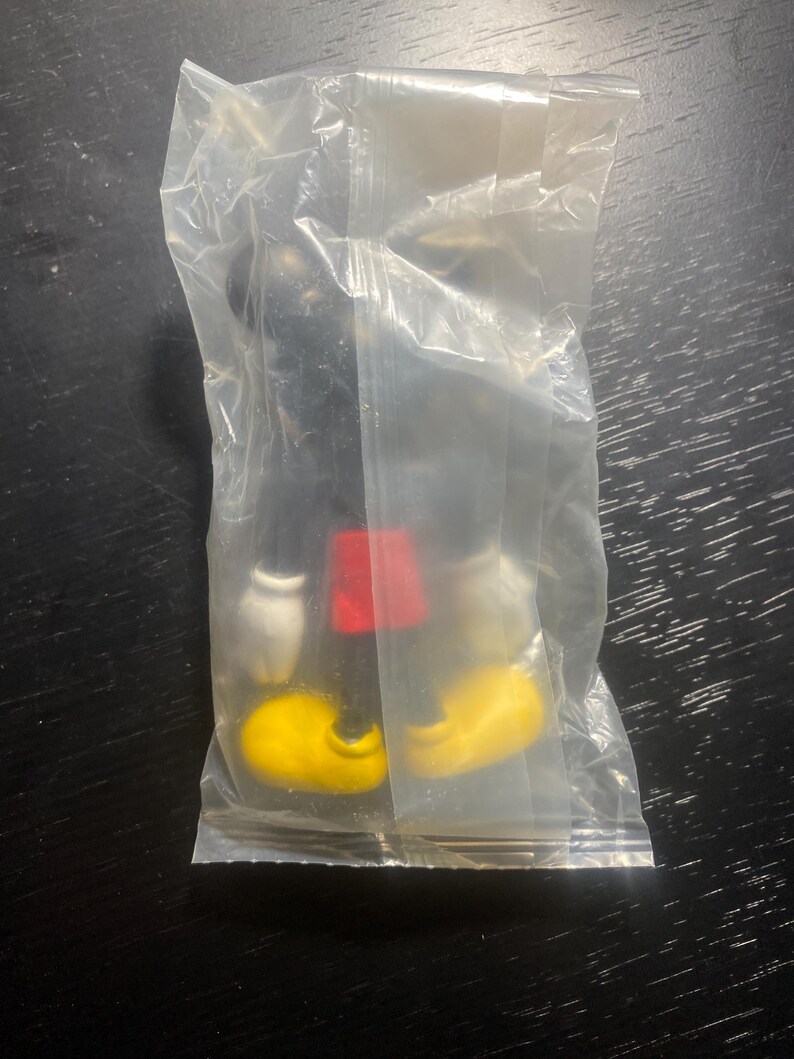 May include: A black Mickey Mouse figurine in a clear plastic bag. The figurine has yellow shoes, white gloves, and red shorts. The figure is on a dark surface.