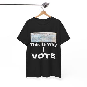 Può includere: T-shirt nera con la scritta bianca "This Is Why I VOTE" sotto un documento d'epoca. Il documento è un "Certificato di Esenzione Fiscale" con dettagli manoscritti. La maglietta è appesa a una gruccia di legno.