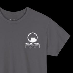 May include: Dark gray t-shirt featuring the Black Mesa logo in white. The logo includes a stylized 'G' shape and the text 'BLACK MESA ANOMALOUS MATERIALS DEPARTMENT'. The shirt also has the text 'S THE GENTLE TEE'.