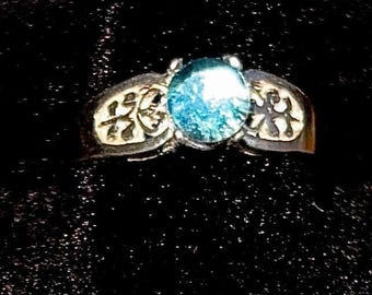 May include: A silver ring featuring a blue gemstone set in the centre. The band is adorned with intricate designs.
