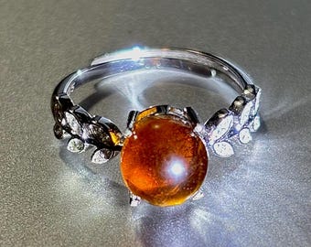 May include: A silver ring featuring a large, round, amber-coloured gemstone. The ring is adorned with small, clear gemstones set in a leaf design.