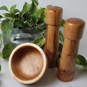 May include: A wooden bowl and two wooden salt and pepper grinders are displayed on a white surface. The grinders are tall and cylindrical, with a rounded top. The bowl is round and shallow. Green leafy plants are in the background.