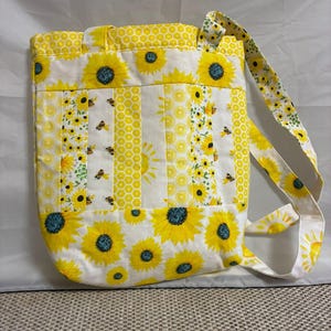 May include: A handmade tote bag with a sunflower and bee pattern. The bag features a white background with yellow sunflowers and bees, and yellow honeycomb and sunburst stripes. The bag has a matching strap and is made of fabric.