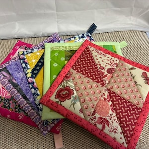 Op de afbeelding: Een verzameling handgemaakte, gewatteerde pannenlappen in verschillende kleuren en patronen. De pannenlappen hebben een mix van bloemen-, geometrische en abstracte ontwerpen, met roze, rode, groene en paarse accenten. Elke pannenlap heeft een lus om op te hangen.