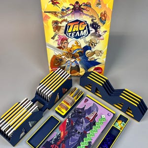 May include: A board game called "TAG TEAM" with a yellow and blue box featuring cartoon characters. The game includes blue and yellow card holders, cards, and game pieces. The "Mordred" card is visible.
