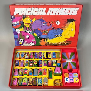 Puede incluir: Juego de mesa "MAGICAL ATHLETE" en una caja roja. Incluye fichas de personajes, dados, cartas y un contenedor de puntos. La ilustración de la caja presenta un plátano con sombrero y otros personajes caprichosos.