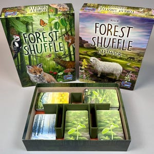 May include: Two board game boxes titled "Forest Shuffle" with nature-themed illustrations. One box features a fox, deer, and butterfly, while the other shows a sheep and landscape. The game box is open, revealing cards and dividers.