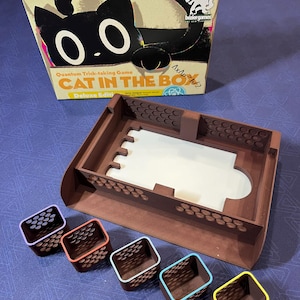 Puede incluir: Una caja marrón con una ilustración de un gato en la parte delantera. La caja está etiquetada como "Quantum Trick-taking Game Cat in the Box Deluxe Edition". La caja está abierta y muestra una bandeja marrón con cinco pequeños contenedores de colores.