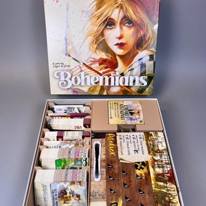 May include: A board game titled "Bohemians" with box art featuring a woman with blonde hair and a red hat. The game box is open, revealing game components, including cards, wooden game pieces, and the text "Atelier".
