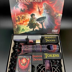 Könnte beinhalten: Geöffnete Spielbox von The Lord of the Rings: Fate of the Fellowship. Das Box-Art zeigt Charaktere und Szenen aus dem Fantasy-Roman. Im Inneren befinden sich Karten, Spielsteine und Spielteile mit der Aufschrift "Friendly Troops" und "Shadow Troops".