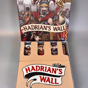 May include: The board game "Hadrian's Wall" is displayed, showing a Roman soldier on the box. The box is open, revealing game pieces and the title. The game's theme is Roman history.