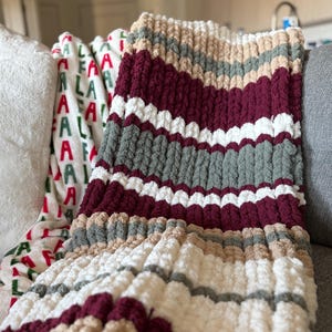 May include: A cozy, hand-knitted blanket in a striped pattern of burgundy, white, gray, and tan. The chunky knit blanket is draped over a sofa, with a white pillow and a festive holiday blanket visible in the background.
