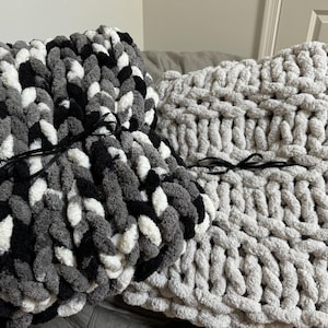 May include: Two chunky knit throw pillows, one in black, white, and gray, and the other in a solid light gray. Both pillows have black ribbon ties.