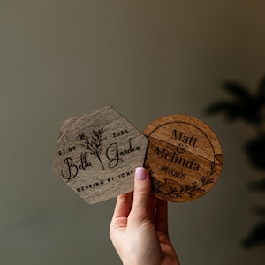 May include: Two wooden wedding coasters. One is a hexagon with "Bella Garden" and the date 2025. The other is round with "Matt & Melinda" and the date 09.13.2025. Both are held up by a person.