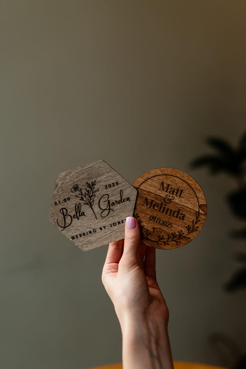 May include: Two wooden wedding coasters. One is a hexagon with "Bella Garden" and the date 2025. The other is round with "Matt & Melinda" and the date 09.13.2025. Both are held up by a person.