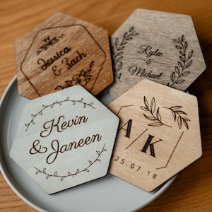 May include: Four hexagonal wooden coasters with engraved names and dates. The coasters are in shades of light and dark brown, with floral and geometric designs. The names "Jessica & Zach", "Rylee & Michael", "Kevin & Janeen", and "A/K 25.07.18" are visible.
