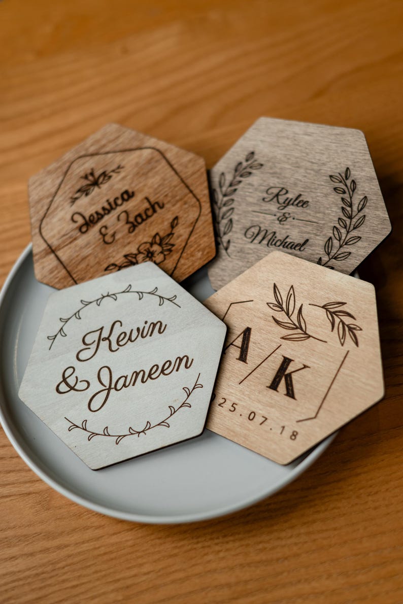 May include: Four hexagonal wooden coasters with engraved names and dates. The coasters are in shades of light and dark brown, with floral and geometric designs. The names "Jessica & Zach", "Rylee & Michael", "Kevin & Janeen", and "A/K 25.07.18" are visible.