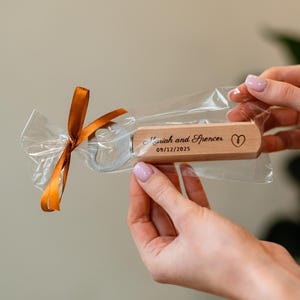 Op de afbeelding: Een houten flesopener met een zilveren openeruiteinde, verpakt in doorzichtig cellofaan en vastgebonden met een oranje lint. De opener is gegraveerd met de namen "Mariah and Spencer", een hartsymbool en de datum "09/12/2025".