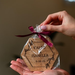 May include: A wooden, hexagon-shaped coaster with the names "CALEB" and "ISABELLA" and the date "5-31-25" engraved on it. The coaster is packaged in clear cellophane and tied with a burgundy ribbon. Floral design accents the names.