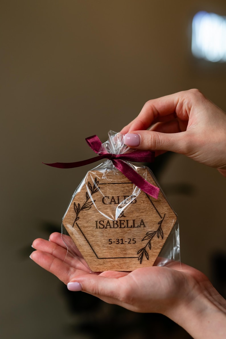 May include: A wooden, hexagon-shaped coaster with the names "CALEB" and "ISABELLA" and the date "5-31-25" engraved on it. The coaster is packaged in clear cellophane and tied with a burgundy ribbon. Floral design accents the names.