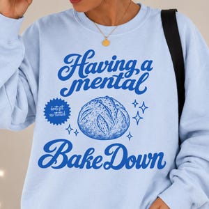 May include: A light blue crewneck sweatshirt featuring dark blue text that reads Having a mental Bake Down. The design includes a graphic of a round loaf of bread, small star icons, and a circular badge with the text don't get me started.