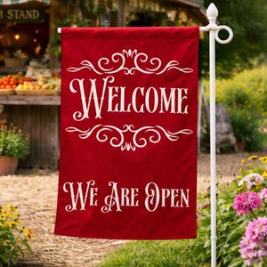 May include: A red outdoor flag with the words "WELCOME WE ARE OPEN" in white script. The flag is displayed on a white pole in a garden setting, with a farm stand in the background.