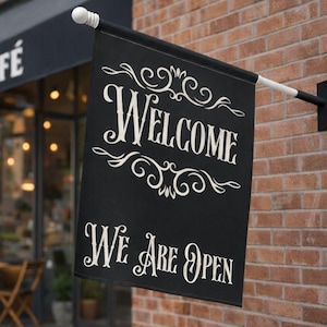 May include: Black rectangular flag with the words "WELCOME" and "WE ARE OPEN" in white script. The flag is hanging on a brick wall outside a cafe.