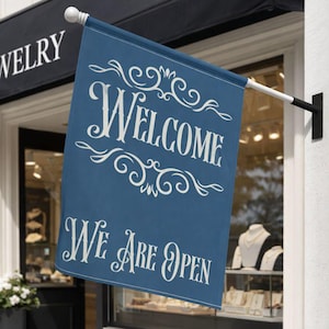 May include: A blue shop flag with the words "WELCOME" and "WE ARE OPEN" in white script. The flag is hanging outside a jewelry store, with the word "JEWELRY" visible on the awning.