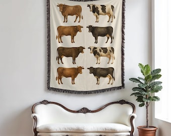 Op de afbeelding: Een crèmekleurige sprei met een herhalend patroon van koeien in aquarel-stijl in verschillende tinten bruin, zwart en wit. De sprei heeft een donkerbruine franje en wordt aan een muur boven een witte bank getoond.