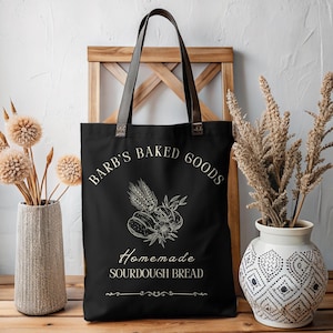 Könnte beinhalten: Schwarze Stofftasche mit weißem Text, der "Barb's Baked Goods" und "Homemade Sourdough Bread" lautet. Die Tasche zeigt eine Strichzeichnung von Brot und Weizen.
