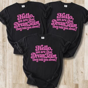 Puede incluir: Tres camisetas negras de manga corta con el texto rosa "Hello, we are that DreamTeam they told you about!" impreso en una fuente negrita y estilizada en la parte delantera. Las camisetas están dispuestas en grupo, mostrando el mismo diseño.
