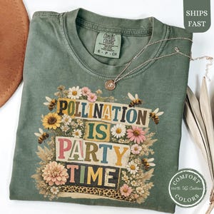 May include: A sage green t-shirt featuring the text "POLLINATION IS PARTY TIME" in colourful block letters, surrounded by bees, sunflowers, and other flowers. A gold necklace rests on the shirt. The shirt is made of 100% US cotton.